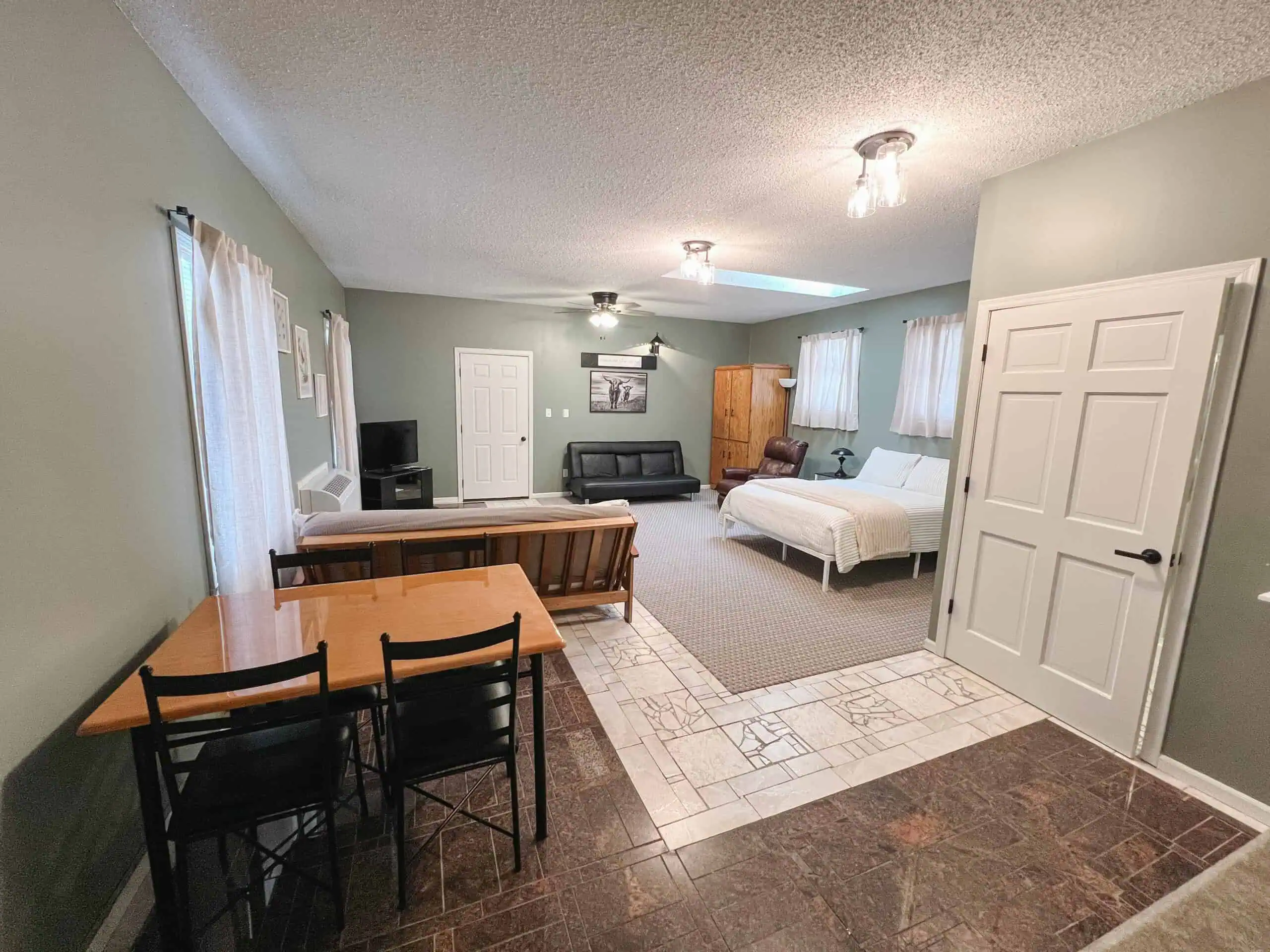 A spacious room with a bed, sofa, dining table, and chairs. The room has light green walls, two windows with curtains, and tiled flooring near the entrance. Ceiling lights provide illumination.