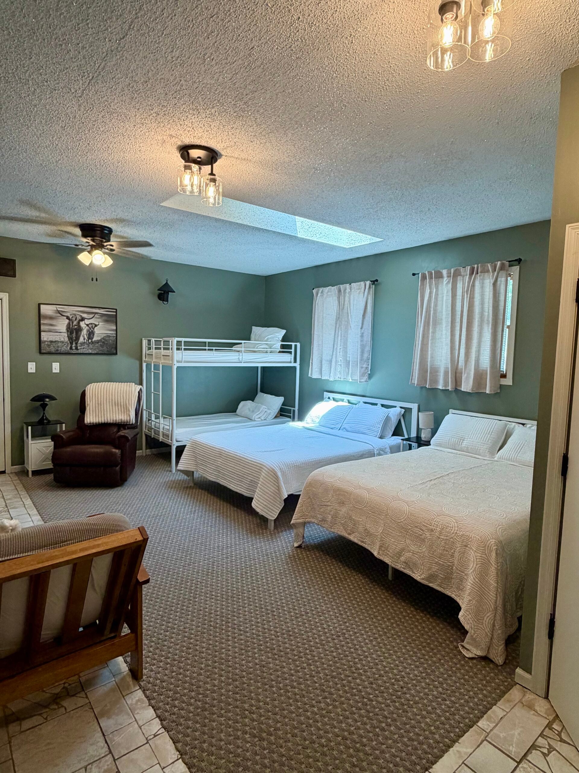 Bedroom with two beds, a bunk bed, a futon, an armchair, and green walls. The room has carpeted flooring, two windows with curtains, and ceiling lights.