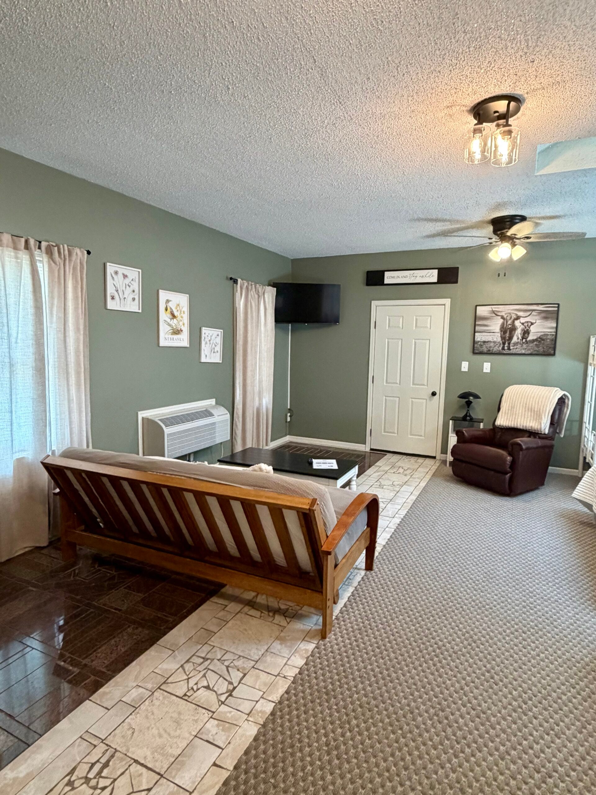 Living room with green walls, futon, armchair, coffee table, wall art, TV, white door, ceiling fan, and tiled entryway. Light from windows with sheer curtains brightens the space.