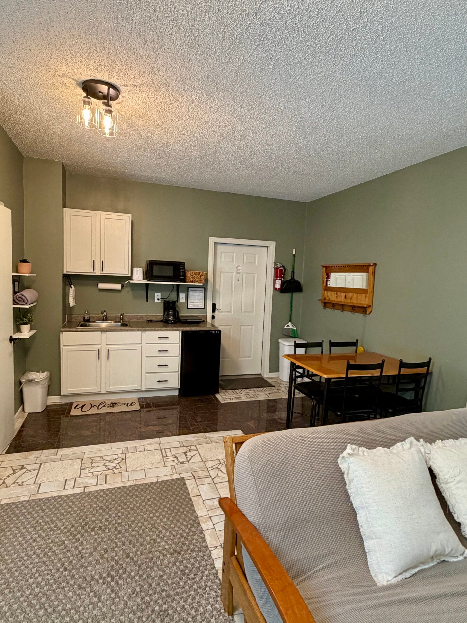 Small kitchen and dining area in a studio apartment with green walls, white cabinets, appliances, a dining table with chairs, and a sofa with white pillows in the foreground.