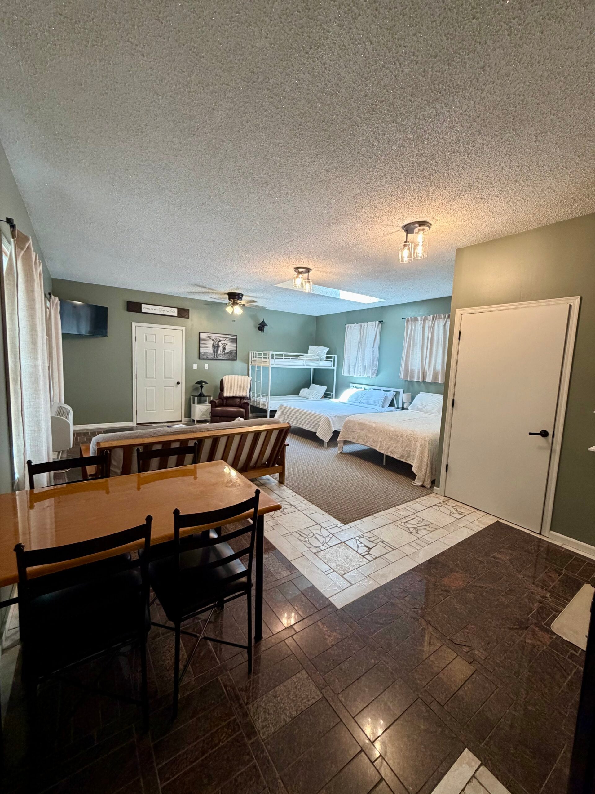Spacious room with two beds, a bunk bed, small dining table with chairs, armchair, TV on the wall, and tiled flooring.