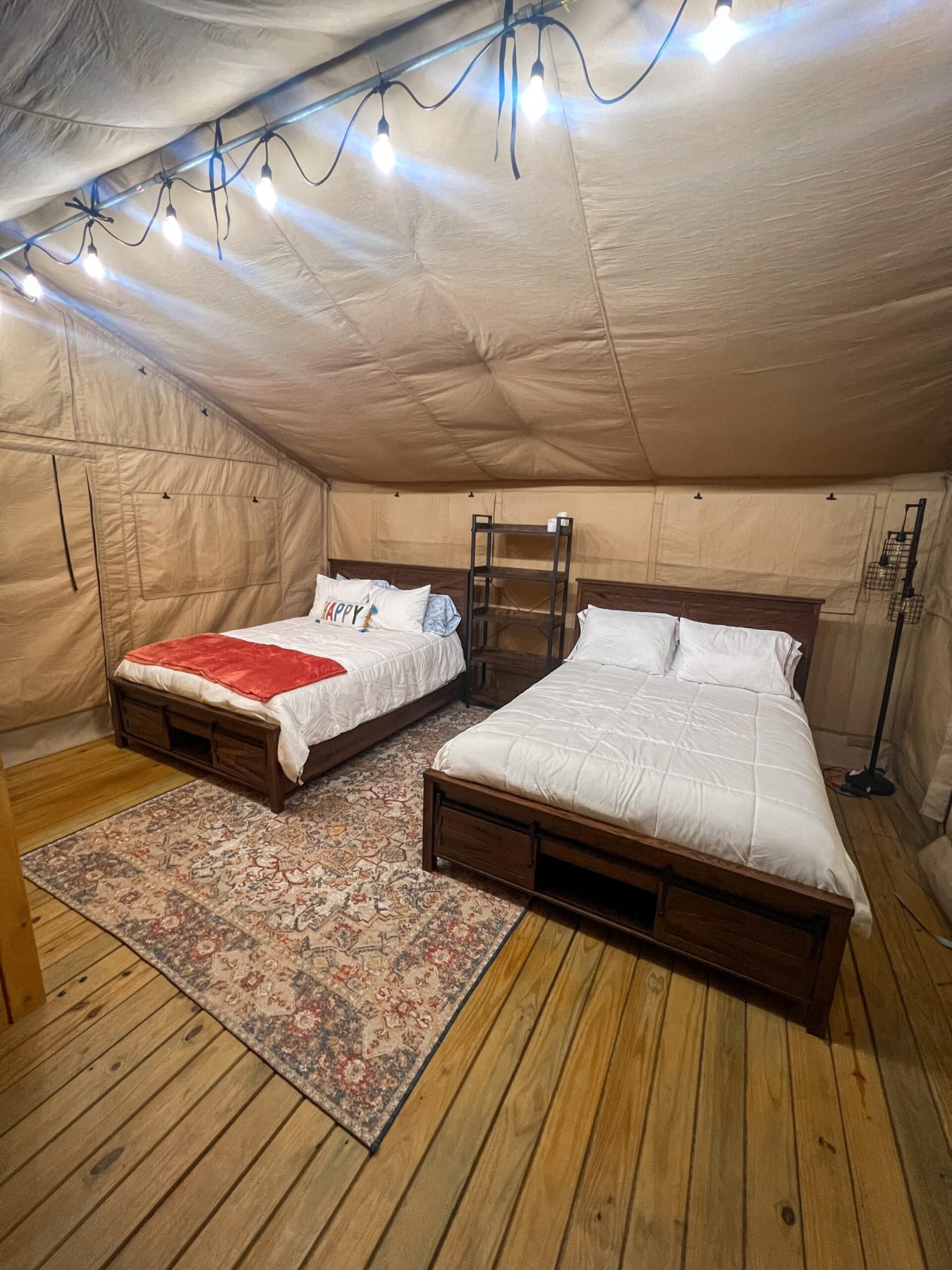 Two double beds with white bedding in a canvas tent, wooden floor, patterned rug, shelving unit, and string lights overhead.